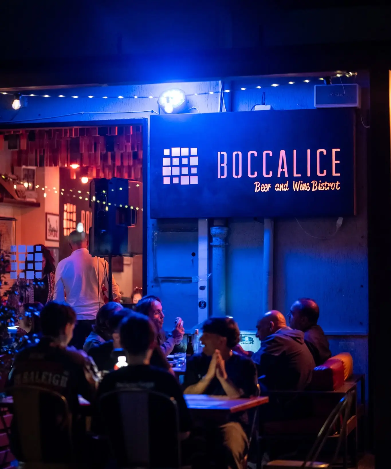 L'ingresso di Boccalice di sera, con l'insegna illuminata di blu e i tavoli all'aperto pieni